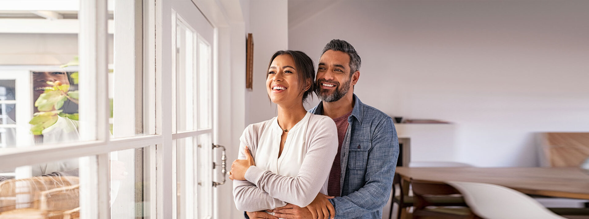 Smiling mid adult couple hugging each other and standing near window while looking outside. Happy and romantic mature man embracing hispanic wife from behind while standing at home with copy space. Future, vision and daydream concept.