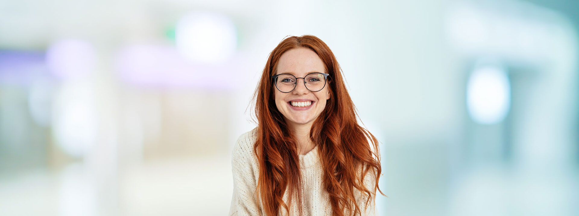 Smiling friendly young woman with long red hair wearing spectacles looking at the camera with a vivacious smile against a white interior wall with copy space