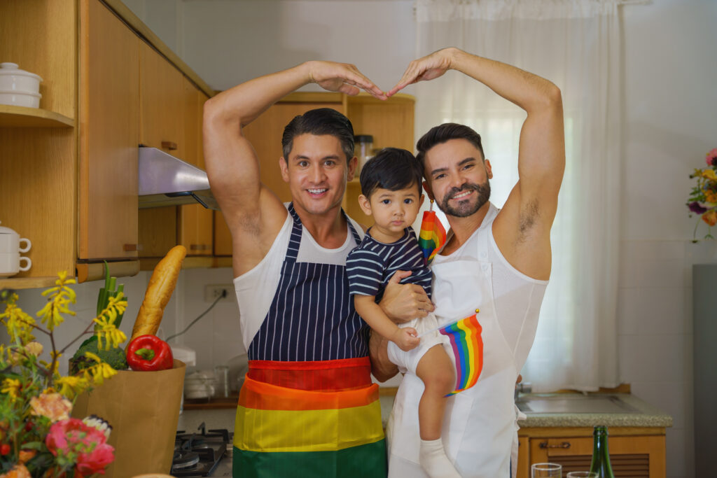 caucasian lgbtq gay couple holding adopted child and enjoying cooking food together in kitchen. concept of LGBTQ same sex couple relationship